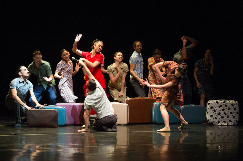 Performers dance as if they were at a party around brightly colored suitcases